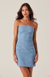 A young woman is wearing a strapless light blue denim dress that is fitted and slightly ruched at the sides. The dress is above knee length and has a simple, sleek design. She is accessorized with gold earrings and a ring on her left hand. Her long, dark brown hair is styled in loose waves.
